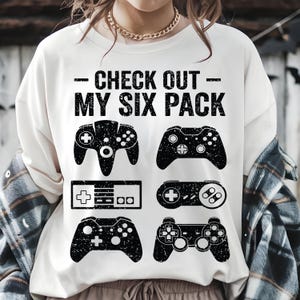 May include: White t-shirt with the text "- CHECK OUT - MY SIX PACK" above six black controller graphics. The controllers are in various shapes and sizes, representing different gaming consoles. A fun and playful design for gamers.