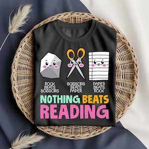 Puede incluir: Camiseta negra con un diseño de Piedra, Papel, Tijeras y gráficos de dibujos animados. El texto "NADA SUPERA LA LECTURA" se muestra en letras coloridas. La camiseta se exhibe en una cesta tejida.