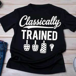 Może przedstawiać: Czarna koszulka z białym napisem "Classically TRAINED", wraz z ilustracjami pedałów samochodowych i dźwigni zmiany biegów. Koszulka ma okrągły dekolt i krótkie rękawy. Projekt znajduje się z przodu.