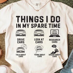 May include: Cream-coloured t-shirt featuring black text and car illustrations. The text says "THINGS I DO IN MY SPARE TIME" with car graphics and phrases such as "DRIVE CARS" and "DREAM ABOUT CARS".