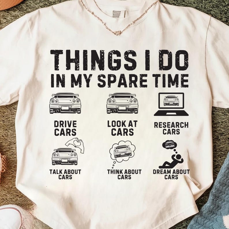 May include: Cream-coloured t-shirt featuring black text and car illustrations. The text says "THINGS I DO IN MY SPARE TIME" with car graphics and phrases such as "DRIVE CARS" and "DREAM ABOUT CARS".