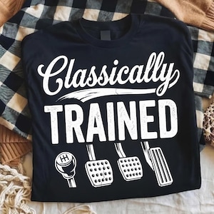 Może przedstawiać: Czarna koszulka z białym napisem "Classically TRAINED", wraz z grafiką dźwigni zmiany biegów i pedałów. Koszulka jest prezentowana na kraciastej narzucie.