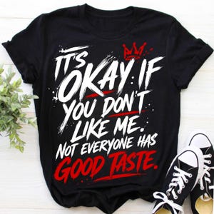 May include: A black short-sleeved t-shirt featuring white and red graphic text that reads: IT'S OKAY IF YOU DON'T LIKE ME. NOT EVERYONE HAS GOOD TASTE. A small red crown icon is positioned above the text, which is styled in a bold, brush-stroke font.