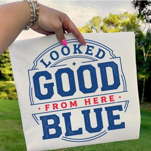 May include: A white t-shirt featuring the text "LOOKED GOOD FROM HERE BLUE" in a blue, vintage design. The text is arranged in a circular and rectangular layout. The shirt is held up by a hand with a bracelet.