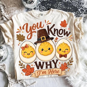 Könnte beinhalten: Ein cremefarbenes T-Shirt mit einer Grafik von drei gefüllten Eiern mit Gesichtern. Das mittlere Ei trägt einen Pilgerhut. Der Text "You know why I'm here" ist in Braun und Orange gehalten, ergänzt durch Herbstblätter und kleine orange Punkte.
