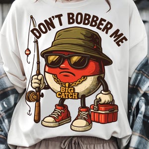 May include: White t-shirt with a cartoon graphic of a red and white fishing bobber wearing sunglasses and a hat, holding a fishing rod and tackle box. Text reads "DON'T BOBBER ME" and "BIG CATCH".