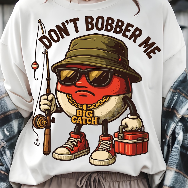 May include: White t-shirt with a cartoon graphic of a red and white fishing bobber wearing sunglasses and a hat, holding a fishing rod and tackle box. Text reads "DON'T BOBBER ME" and "BIG CATCH".