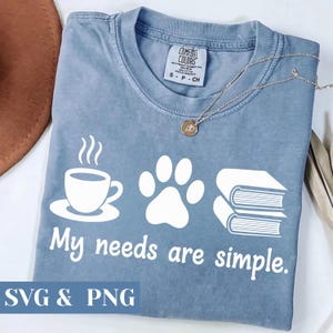 May include: Blue Comfort Colors t-shirt with a white graphic of a coffee cup, paw print, and books. The text "My needs are simple" is below the graphics. A silver necklace with a pendant is on the shirt.