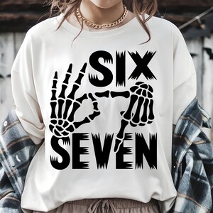 May include: Cream-colored sweatshirt with a black graphic design. The design features the word "SIX" above the word "SEVEN", with skeletal hands forming the numbers. The sweatshirt is a casual, long-sleeved top.
