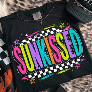 以下が含まれることがあります： 鮮やかなネオンピンク、ライムグリーン、オレンジ、ティールの文字で「SUNKISSED」と書かれた黒のTシャツ。デザインには白黒の市松模様の背景、カラフルな星のアクセント、首元にComfort Colorsのブランドラベルが含まれています。
