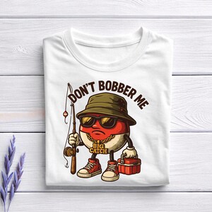 May include: White t-shirt featuring a cartoon character with a fishing rod, sunglasses, and a hat. The character has a red and white body and is holding a red tackle box. Text on the shirt reads "DON'T BOBBER ME" and "BIG CATCH".