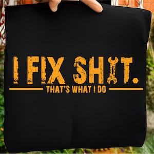 Op de afbeelding: Zwarte draagtas met de tekst "I FIX SHIT. THAT'S WHAT I DO" in verweerde oranje letters. Het ontwerp bevat een moersleutelafbeelding. De tas wordt door een hand omhoog gehouden.