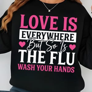 May include: Black sweatshirt with the text "LOVE IS EVERYWHERE But So Is THE FLU WASH YOUR HANDS" in pink and white. The design includes pink hearts. The sweatshirt is a casual garment.