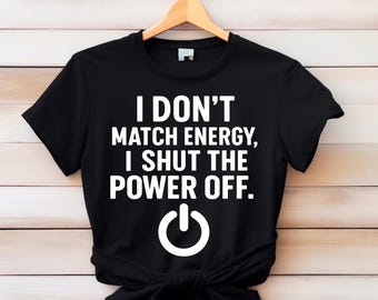 Könnte beinhalten: Schwarzes T-Shirt mit dem weißen Text "I DON'T MATCH ENERGY, I SHUT THE POWER OFF." und einem weißen Power-Button-Symbol. Das Shirt hängt an einem Holzbügel vor einem hellen Holz Hintergrund.