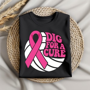 May include: Black t-shirt featuring a pink ribbon and the words "DIG FOR A CURE" in pink. The design includes a white circle with curved lines behind the ribbon and text. The shirt is folded and placed on a woven surface.