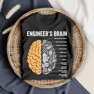 Może przedstawiać: Czarny t-shirt z grafiką mózgu inżyniera, podzielonego na dwie połowy. Jedna strona to stylizowany mózg, druga to schemat mechaniczny. Tekst zawiera "Engineer's Brain" i humorystyczne etykiety.