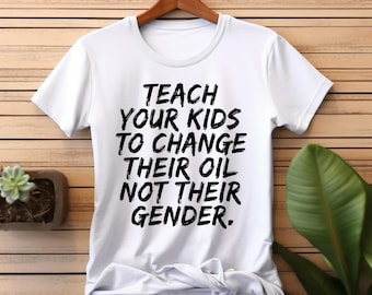 Op de afbeelding: Wit T-shirt met de tekst "TEACH YOUR KIDS TO CHANGE THEIR OIL NOT THEIR GENDER." De tekst is vetgedrukt en zwart. Het shirt hangt aan een houten hanger tegen een houten achtergrond.