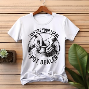 May include: White t-shirt with a black graphic of hands shaping a pot on a pottery wheel. The text "SUPPORT YOUR LOCAL POT DEALER" is printed above and below the graphic in a circular design. The shirt is hanging on a wooden hanger.
