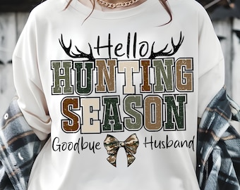 Puede incluir: Sudadera blanca de manga larga con el texto "Hello Hunting Season Goodbye Husband" en una mezcla de fuentes y colores. El diseño incluye astas y un lazo de camuflaje. La sudadera es de estilo casual.