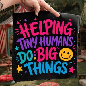May include: A black fabric square featuring the colorful text HELPING TINY HUMANS DO BIG THINGS in pink, purple, and blue lettering. The design includes a yellow smiley face icon and various small star shapes in pink, purple, and yellow colors.