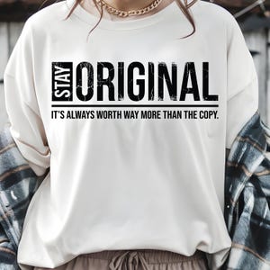 May include: White long-sleeve shirt with the words "STAY ORIGINAL" in bold black letters. Below the text is the phrase "IT'S ALWAYS WORTH WAY MORE THAN THE COPY." The shirt is a casual style.