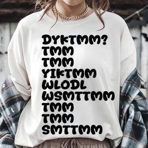 May include: Cream-colored sweatshirt with the text "DYKTMM? TMM TMM YIKTMM WLODL WSMTTMM TMM TMM SMTTMM" in bold black letters. The shirt is a casual style.