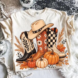 Könnte beinhalten: Ein cremefarbenes T-Shirt mit einer grafischen Illustration von drei Cowboystiefeln, einem beigen Cowboyhut, Kürbissen und Herbstblättern. Die Stiefel zeigen schwarze Blattmuster, weiße Gänseblümchen sowie ein schwarz-weißes Karomuster.