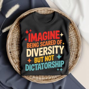 May include: Black t-shirt with the text "IMAGINE being scared of DIVERSITY but not DICTATORSHIP" in red, orange, yellow, and blue. The shirt is displayed in a woven basket.