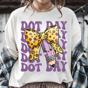 May include: Cream-colored sweatshirt featuring a graphic of a purple pencil with a yellow polka-dot bow and the text "DOT DAY" in purple. The pencil has a smiling face. A gold chain is visible.