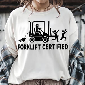 Może przedstawiać: Biała koszulka z czarną grafiką wózka widłowego, postacią z patyczaków, która go prowadzi, i dwiema postaciami z patyczaków uciekającymi. Pod grafiką znajduje się napis "FORKLIFT CERTIFIED".