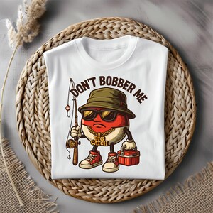 May include: White t-shirt with a cartoon character wearing sunglasses, a fishing hat, and holding a fishing rod. The text "DON'T BOBBER ME" is printed above the character, and "BIG CATCH" is printed below.