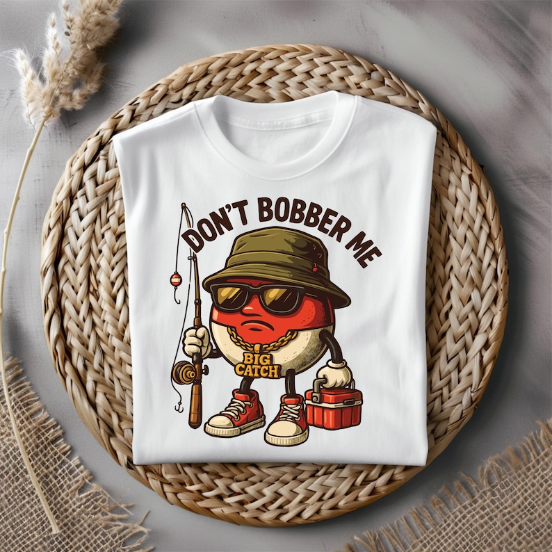 May include: White t-shirt with a cartoon character wearing sunglasses, a fishing hat, and holding a fishing rod. The text "DON'T BOBBER ME" is printed above the character, and "BIG CATCH" is printed below.