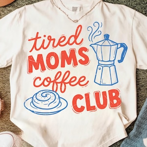 Può includere: T-shirt bianco con le parole "tired MOMS coffee CLUB" in rosso e blu. Il design include una caffettiera blu, un rotolo di cannella e una collana. La maglietta è realizzata in materiale morbido.