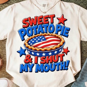 May include: A white t-shirt featuring a graphic of a pie with a crust and a filling patterned like the American flag. The text reads SWEET POTATO PIE & I SHUT MY MOUTH! in red and blue letters, accented by red and blue stars on a white background.
