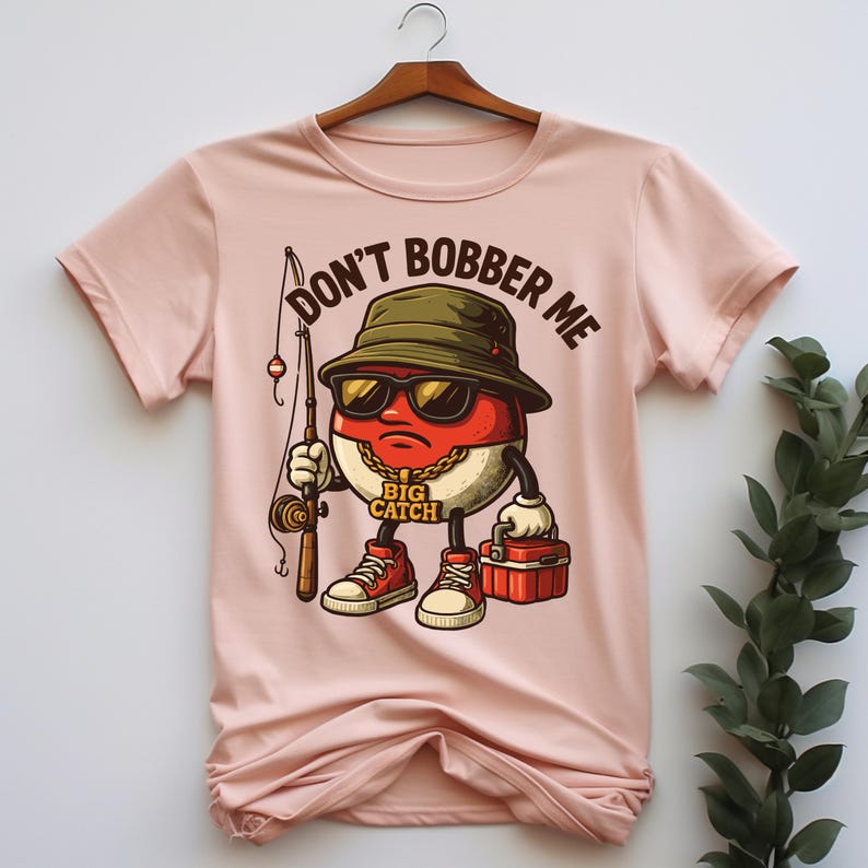 May include: Light pink t-shirt featuring a cartoon character with sunglasses, a fishing rod, and a tackle box. The character is a red and white oval with a gold chain and red sneakers. Text on the shirt reads "DON'T BOBBER ME" and "BIG CATCH".