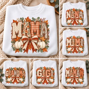 May include: A white crewneck sweatshirt featuring a graphic print of autumn leaves, white pumpkins, and a large bow. The text MAMA, NANA, MIMI, GRANDMA, GIGI, or MAMA W is displayed in a patterned font surrounded by floral and seasonal foliage.