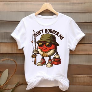 May include: White t-shirt featuring a cartoon character with sunglasses, a fishing rod, and a tackle box. The character is a red and white oval with a gold chain and red sneakers. Text on the shirt reads "DON'T BOBBER ME" and "BIG CATCH".