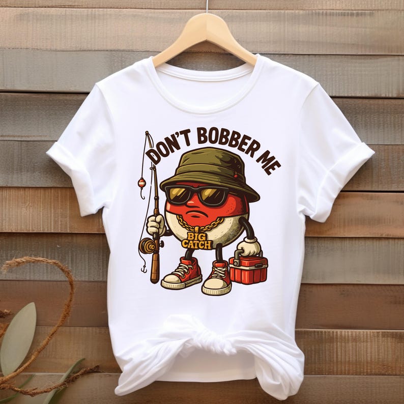May include: White t-shirt featuring a cartoon character with sunglasses, a fishing rod, and a tackle box. The character is a red and white oval with a gold chain and red sneakers. Text on the shirt reads "DON'T BOBBER ME" and "BIG CATCH".
