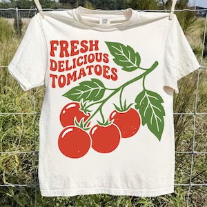 Peut inclure: T-shirt crème avec l'inscription "FRESH DELICIOUS TOMATOES" en rouge, et une illustration verte de tomates et de feuilles. Le t-shirt est suspendu à une corde à linge sur un fond extérieur flou.
