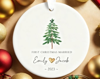 May include: A white ceramic Christmas ornament with a watercolor evergreen tree design. The text reads "First Christmas Married" with the names "Emily" and "Jacob" and the year "2023". A white ribbon is attached for hanging.