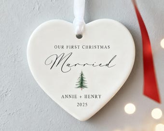 May include: A heart-shaped, white ceramic Christmas ornament with the text "OUR FIRST CHRISTMAS Married" in black script. A small green Christmas tree graphic is below the word "Married". The names "ANNIE + HENRY" and the year "2025" are also printed on the ornament.