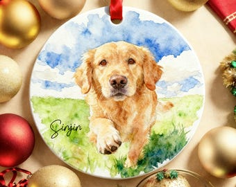 May include: Circular ceramic ornament with a watercolour painting of a golden retriever dog. The dog is in a grassy field with a blue sky. The name "Sinjin" is written in cursive. A red ribbon is attached for hanging.