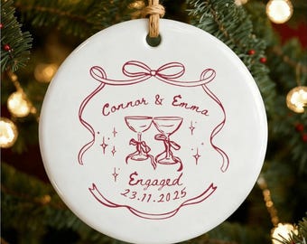 以下が含まれることがあります： 白のセラミック製クリスマスオーナメント。マルーンのデザインで、「Connor & Emma」の名前、2つのシャンパングラス、「Engaged」の文字と日付23.11.2025が描かれています。上部には吊り下げ用の紐ループが付いています。