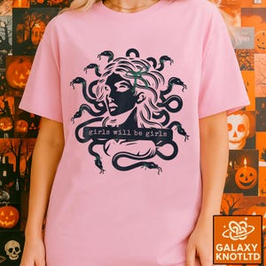 Puede incluir: Camiseta rosa claro con un gr&aacute;fico negro de Medusa y el texto "girls will be girls". El dise&ntilde;o de Medusa presenta serpientes como cabello. La camiseta tiene mangas cortas y cuello redondo.