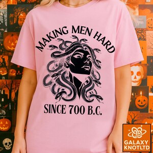May include: A light pink t-shirt featuring a black and white graphic of Medusa with snakes for hair. The text "MAKING MEN HARD SINCE 700 B.C." is printed above and below the graphic. The shirt has short sleeves.