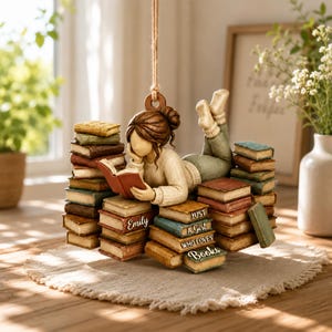 May include: A hanging ornament featuring a figure with brown hair in a bun, wearing a cream sweater and green pants, lying on a stack of books. The books are various colors and include the text "Emily Just A Girl WHO LOVES Books".