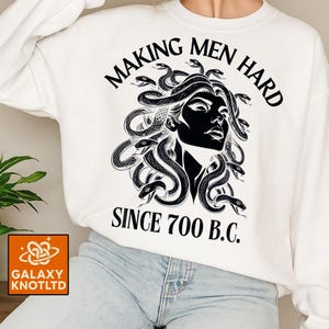 May include: White sweatshirt featuring a black graphic of Medusa with snakes for hair. The text "MAKING MEN HARD SINCE 700 B.C." is printed above and below the image. The sweatshirt has a crew neck.