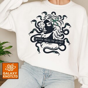 Puede incluir: Sudadera blanca con un gr&aacute;fico negro de Medusa con serpientes como pelo. El texto "girls will be girls" est&aacute; debajo del gr&aacute;fico. La sudadera tiene cuello redondo y mangas largas. El logotipo de Galaxy Knotltd est&aacute; en la esquina inferior izquierda.