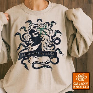 Puede incluir: Sudadera beige claro con un gr&aacute;fico de Medusa, serpientes y el texto "girls will be girls" en azul oscuro. La sudadera tiene cuello redondo y mangas largas.