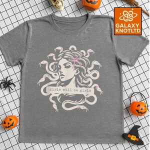 Puede incluir: Camiseta gris con un gr&aacute;fico de Medusa, serpientes y el texto "girls will be girls". El dise&ntilde;o es blanco y rosa. La camiseta tiene cuello redondo y mangas cortas.